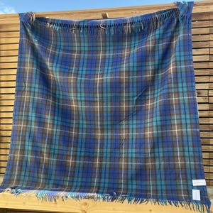Op de afbeelding: Een blauwe, turquoise en grijze geruite deken met franjes langs de onderrand. De deken is over een houten constructie gedrapeerd, waarbij het patroon en de textuur worden benadrukt. Het ruitpatroon heeft een mix van blauwe en turquoise tinten met grijze en witte accenten.