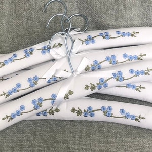 Puede incluir: Un conjunto de perchas acolchadas blancas con un patrón bordado de flores azules y tallos verdes. Cada percha incluye un gancho de metal plateado y un lazo de satén blanco atado en la base del gancho para colgar prendas de vestir.