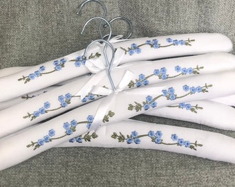Op de afbeelding: Een set witte gevoerde kledinghangers met een geborduurd patroon van blauwe bloemen met groene stelen. Elke hanger is voorzien van een zilverkleurige metalen haak en een wit satijnen lint gestrikt aan de basis van de haak voor het ophangen van kleding.