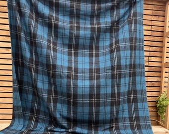 Puede incluir: Una manta a cuadros azul y negro con líneas blancas y flecos. La manta está sobre una estructura de madera, mostrando su patrón y textura. Un elemento de decoración hogareña.