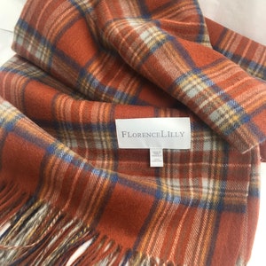Puede incluir: Una manta de lana a cuadros marrones, azules y beige con flecos. La manta está doblada y tiene una etiqueta que dice "Florence Lilly".