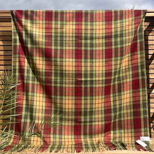 Op de afbeelding: Een grote, rechthoekige geruite deken in tinten rood, groen en geel. De deken hangt en toont een klassiek tartanpatroon. De franje langs de onderrand voegt een decoratief tintje toe. De deken is gemaakt van een zacht materiaal.