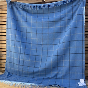 May include: A blue plaid blanket with fringe along the edges. The blanket has a grid pattern in black and tan. The blanket is hanging on a wooden structure. The blanket is made of a soft material.