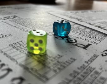 May include: Two translucent six-sided dice, one lime green and one cyan blue, resting on a printed paper game sheet. The dice feature white pips on each face. The game sheet contains columns of text including words like grey, black, red, and purple.