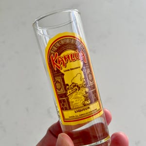 May include: A clear glass shot glass with a yellow and brown Kahlúa liqueur label. The label features the brand name in a stylized font, along with the word "Liqueur" and other text. The glass is cylindrical and appears to be empty.