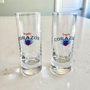 May include: Two clear shot glasses with the words "Tequila CORAZÓN" printed in blue and white, with a red heart and blue flame design. The shot glasses are cylindrical and sit on a light-colored surface.