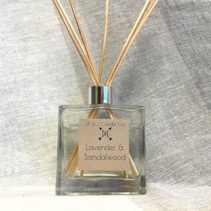 May include: A square glass diffuser bottle with wooden reeds. The label reads "Lavender & Sandalwood" and "EP & J Candle Co." The bottle has a silver cap and is filled with a clear liquid. The background is a light gray fabric.