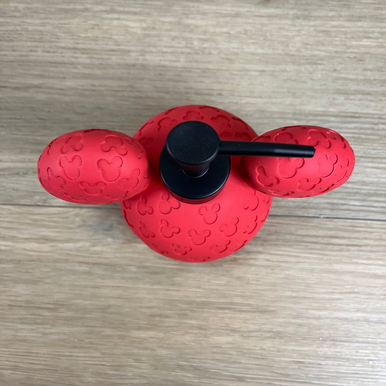 May include: Red soap dispenser shaped like Mickey Mouse's head with black pump and Mickey Mouse ears.