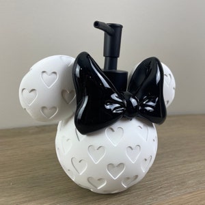 May include: White ceramic soap dispenser shaped like Mickey Mouse's girlfriend, Minnie Mouse, with a black bow and a black pump. The dispenser is covered in a pattern of white hearts.