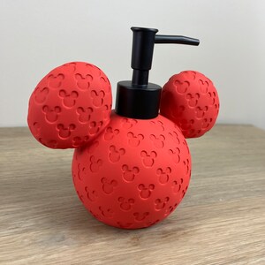 May include: Red soap dispenser shaped like Mickey Mouse's head with black pump and ears. The dispenser is covered in a pattern of small Mickey Mouse heads.