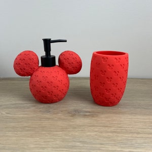 May include: Red Mickey Mouse themed bathroom set with a soap dispenser and a tumbler. The dispenser is shaped like Mickey Mouse's head with black ears and a pump.