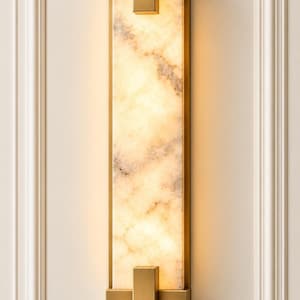 May include: A rectangular wall sconce featuring a vertical slab of translucent, cream-colored stone with grey veining, encased in a brushed brass metal frame with geometric brackets at the top and bottom that emit a warm, soft glow.