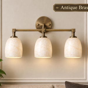 May include: A three-light wall fixture with an antique brass finish and a circular mounting plate. The fixture features three bell-shaped, cream-colored marbled glass shades. A rectangular label in the top right corner reads Antique Brass.