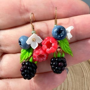 May include: A pair of gold earrings with a cluster of polymer clay berries. The earrings feature red raspberries, blackberries, and blueberries, as well as white flowers.
