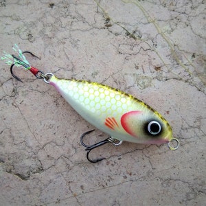Puede incluir: Un señuelo de pesca amarillo y blanco con una aleta roja y naranja y un ojo negro. El señuelo tiene una cola de plumas verde y blanca y tres anzuelos negros.