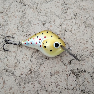Puede incluir: Un señuelo de pesca dorado y blanco con manchas negras y rojas. El señuelo tiene un solo anzuelo y un labio plateado.