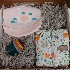 May include: A gift box filled with baby items, including a pink silicone bib with a swan design, a stackable set of colorful silicone cups, and a white muslin swaddle blanket with a woodland animal print.