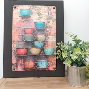 May include: A framed wall art piece featuring a stack of colorful teacups and saucers against a brick wall background. The teacups are in shades of blue, green, and red, and the saucers are in shades of brown and white.