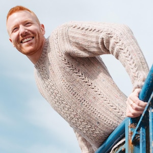 Può includere: Un maglione lavorato a maglia beige strutturato con dettagli a treccia e punti strutturati. Il maglione ha un girocollo e maniche lunghe. La persona sorride e si appoggia a una ringhiera blu. Il cielo è blu.