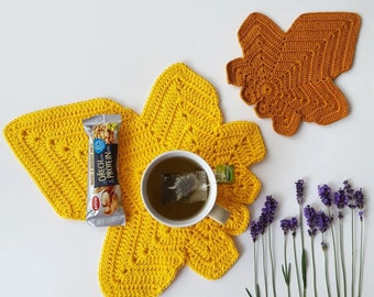 May include: A large yellow crochet leaf-shaped mat, a smaller orange crochet leaf-shaped mat, a white mug with tea, an Emco nut and protein bar, and several sprigs of purple lavender flowers arranged on a plain white surface.