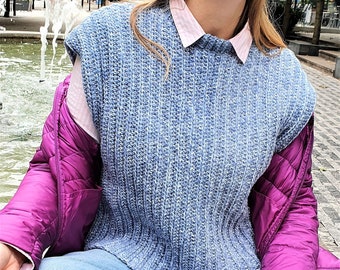 Könnte beinhalten: Eine Frau trägt eine blaue Strickweste über einem weißen Hemd, eine violette Steppjacke und eine blaue Jeans. Sie sitzt mit gekreuzten Beinen und den Händen im Schoß.
