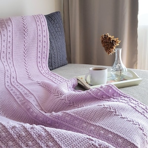 Puede incluir: Una manta de ganchillo lila con patrones texturizados y un cojín a juego. La manta está sobre un sofá, con una bandeja con una taza de café y un jarrón con flores secas.