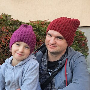 Puede incluir: Primer plano de un padre y un hijo que llevan gorros de punto a juego. El padre lleva un gorro rojo y una chaqueta gris. El niño lleva un gorro morado y una sudadera gris.