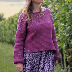Può includere: Una donna indossa un maglione viola con una texture scintillante e una gonna con stampa floreale. È in piedi in un vigneto con viti verdi e una vista lontana di un lago.