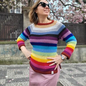 Może przedstawiać: Kobieta w kolorowym, paskowanym swetrze na szydełku z długimi rękawami. Sweter ma wzór tęczy z paskami w kolorze czerwonym, pomarańczowym, żółtym, zielonym, niebieskim, indygo i fioletowym. Kobieta ma na sobie różową spódnicę i związane włosy.