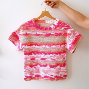 May include: A pink, orange, and white striped crochet top with short sleeves. The top is hanging on a wooden hanger.