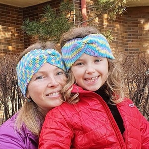 May include: A woman and a young girl wearing colorful knitted headbands. The woman is wearing a purple jacket and the girl is wearing a red jacket.