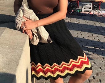 May include: A brown tank top and a black knee-length skirt with a chevron pattern in red, orange, and yellow at the hem. The outfit includes light-colored sneakers and a light-colored patterned jacket held in one hand, with silver sunglasses.