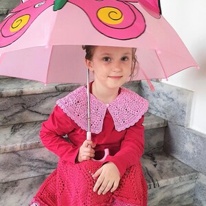 May include: A young girl wearing a red crocheted dress and a pink crocheted collar sits on stairs holding a pink butterfly umbrella.