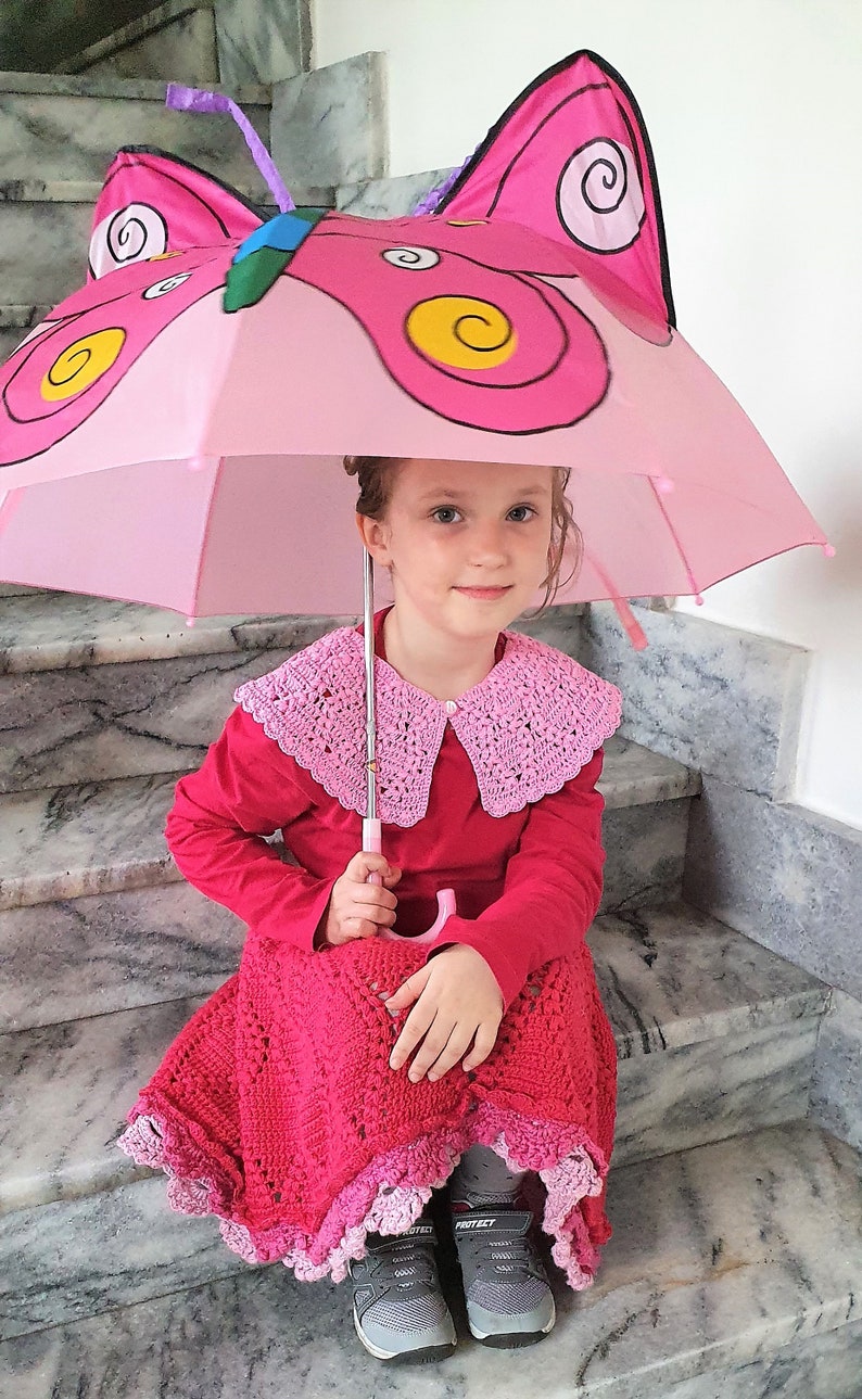 May include: A young girl wearing a red crocheted dress and a pink crocheted collar sits on stairs holding a pink butterfly umbrella.