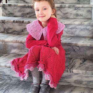 May include: A young girl wearing a pink crocheted collar and a red crocheted skirt with a pink trim. She is sitting on a set of stairs.