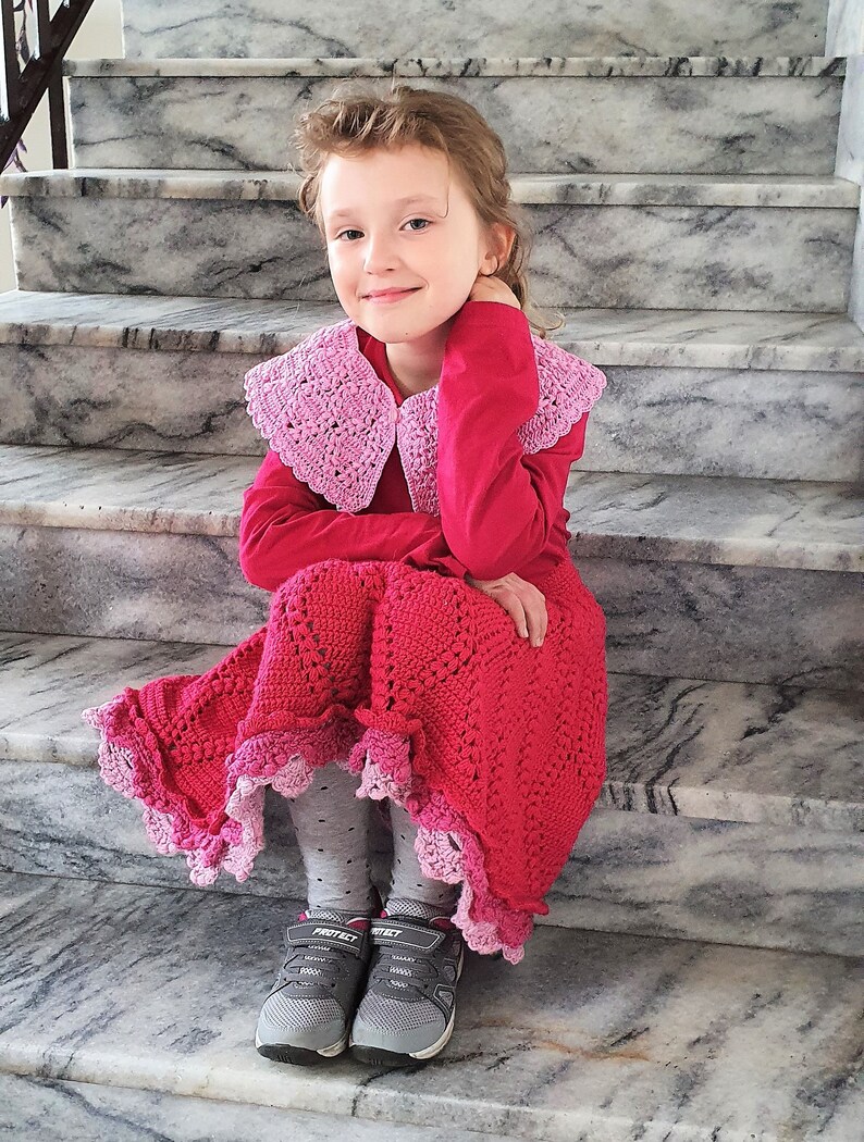 May include: A young girl wearing a pink crocheted collar and a red crocheted skirt with a pink trim. She is sitting on a set of stairs.