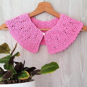 May include: A pink crocheted collar with a pearl button in the centre. The collar is hanging on a wooden hanger against a white wood background.