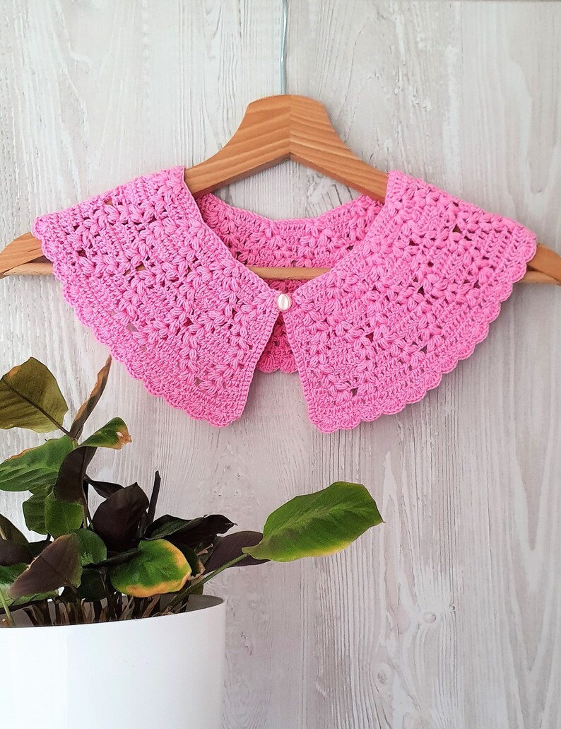 May include: A pink crocheted collar with a pearl button in the centre. The collar is hanging on a wooden hanger against a white wood background.