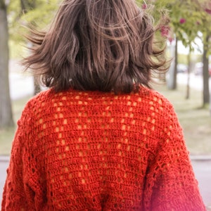 May include: A woman wearing a red crocheted sweater with a black belt. The sweater has a loose fit and a delicate open-stitch pattern.