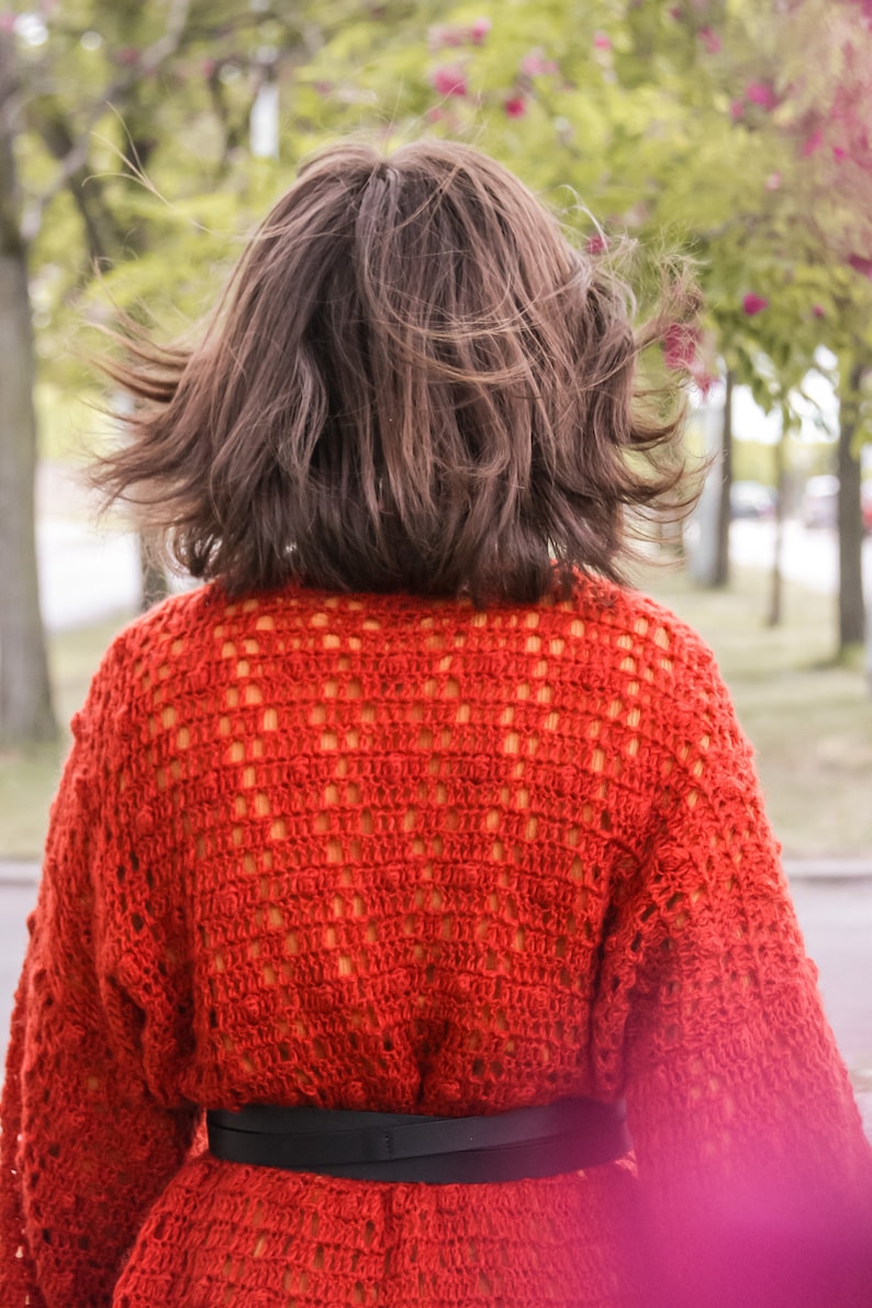 May include: A woman wearing a red crocheted sweater with a black belt. The sweater has a loose fit and a delicate open-stitch pattern.