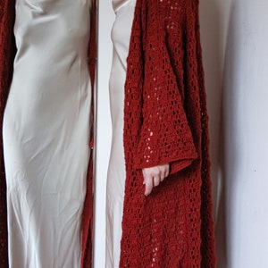 May include: A red crocheted cardigan with a lacy pattern. The cardigan is open at the front and has long sleeves. The cardigan is worn over a white slip dress.