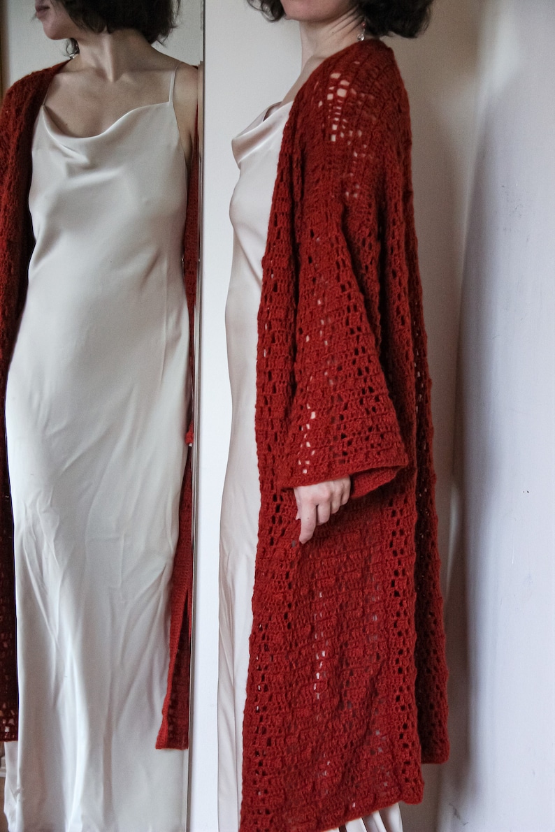 May include: A red crocheted cardigan with a lacy pattern. The cardigan is open at the front and has long sleeves. The cardigan is worn over a white slip dress.