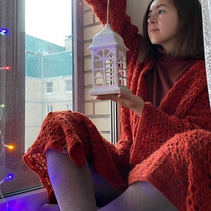 May include: A woman wearing a red crocheted cardigan holds a white ceramic lantern with a decorative design. The lantern is hanging from a string. The woman is sitting by a window with a string of colourful lights.