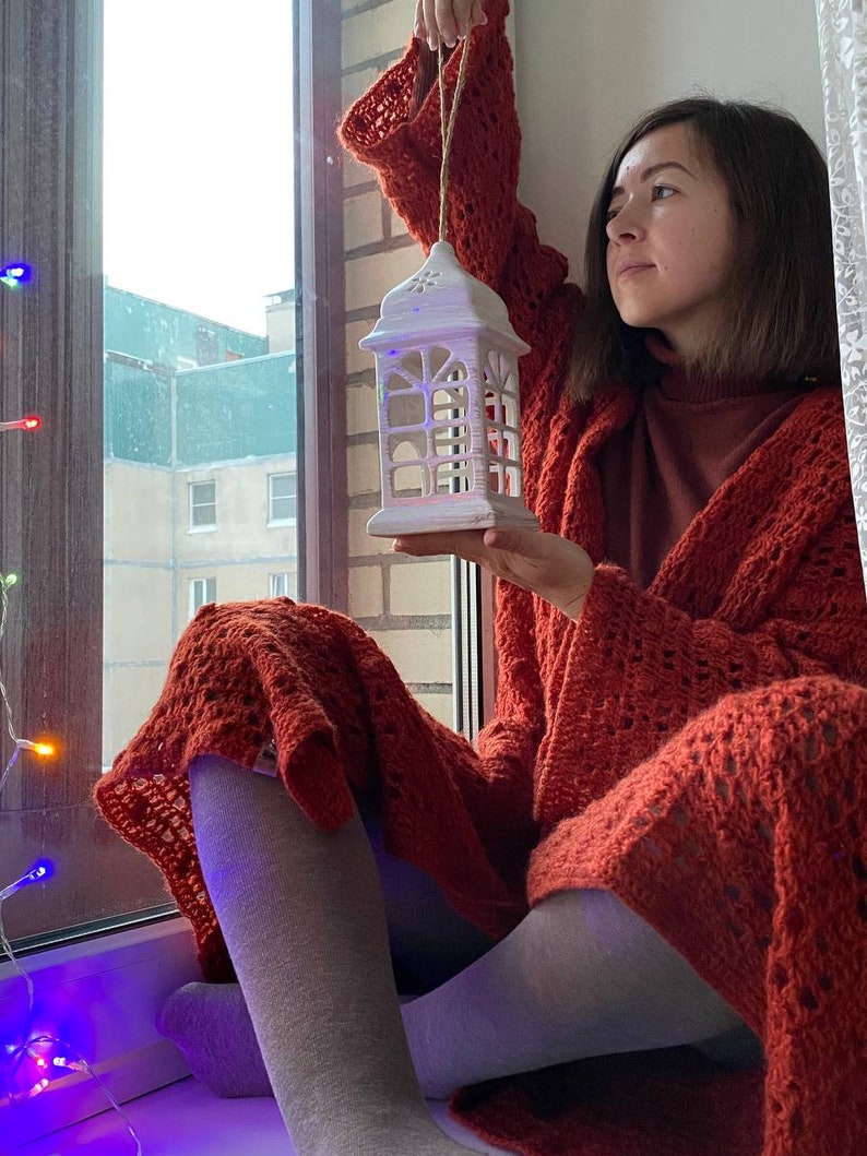 May include: A woman wearing a red crocheted cardigan holds a white ceramic lantern with a decorative design. The lantern is hanging from a string. The woman is sitting by a window with a string of colourful lights.