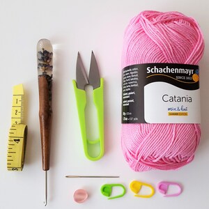 May include: A pink ball of yarn labelled "Schachenmayr Catania mix & knit Summer Edition" with a crochet hook, scissors, a measuring tape, a needle, stitch markers, and a wooden dowel with a clear resin top containing dried lavender flowers.