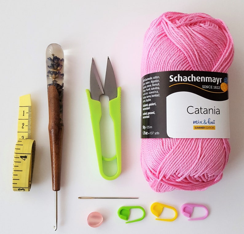 May include: A pink ball of yarn labelled "Schachenmayr Catania mix & knit Summer Edition" with a crochet hook, scissors, a measuring tape, a needle, stitch markers, and a wooden dowel with a clear resin top containing dried lavender flowers.