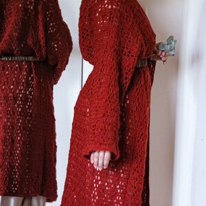 May include: A long, rust-coloured crocheted cardigan with a loose fit and long sleeves. The cardigan is belted at the waist with a brown leather belt.