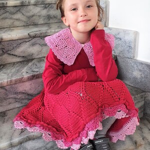 May include: A young girl wearing a red long-sleeved shirt with a pink crocheted collar and a pink and red crocheted skirt. She is sitting on a set of stairs and wearing grey trainers with the word "Protect" on them.