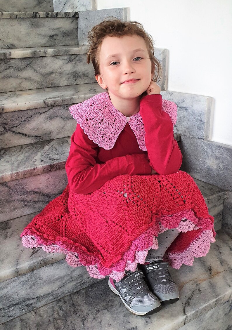 May include: A young girl wearing a red long-sleeved shirt with a pink crocheted collar and a pink and red crocheted skirt. She is sitting on a set of stairs and wearing grey trainers with the word "Protect" on them.