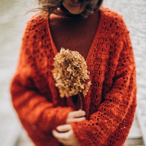 May include: A person wearing a rust-colored, open-weave cardigan over a matching turtleneck. The cardigan has a deep V-neck and long sleeves. The person is holding a dried, beige flower bouquet. The background is blurred, with water and a wooden dock visible.